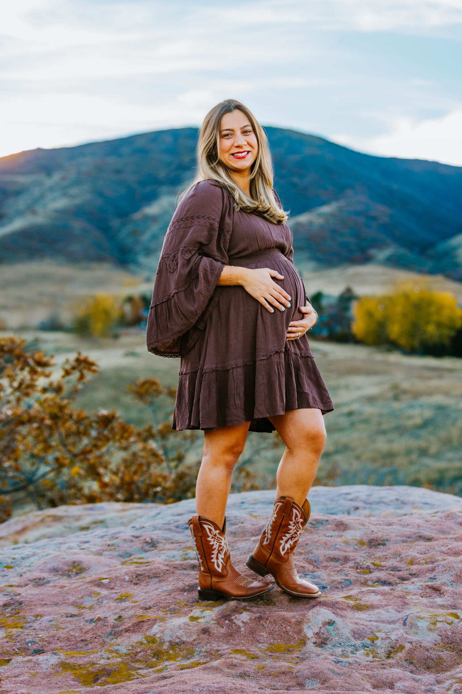 Pregnant woman in brown dress and cowboy boots stands on a rock with mountain scenery backdrop.
