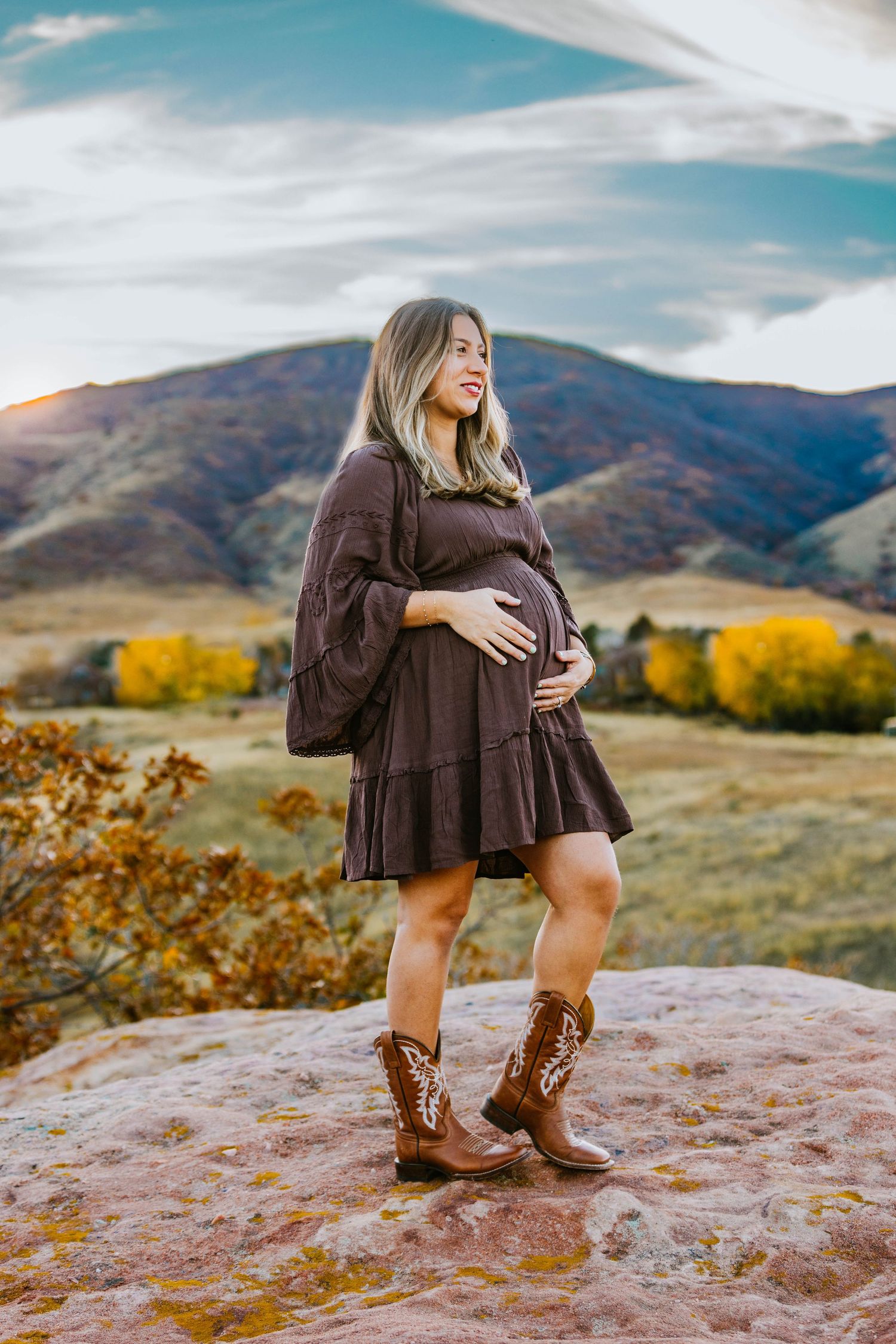 Pregnant woman in brown dress and cowboy boots standing on rocks with autumn mountains backdrop.
