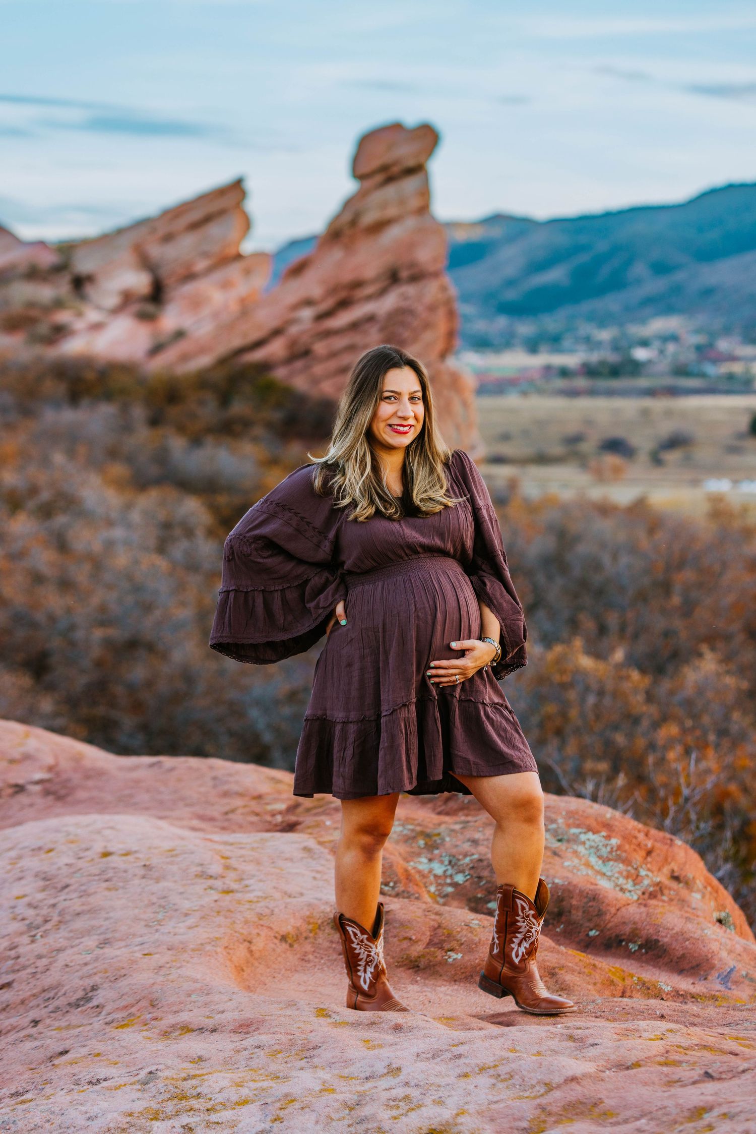 Pregnant woman in burgundy dress and cowboy boots posing at Red Rocks Park, Colorado, cradling baby bump.