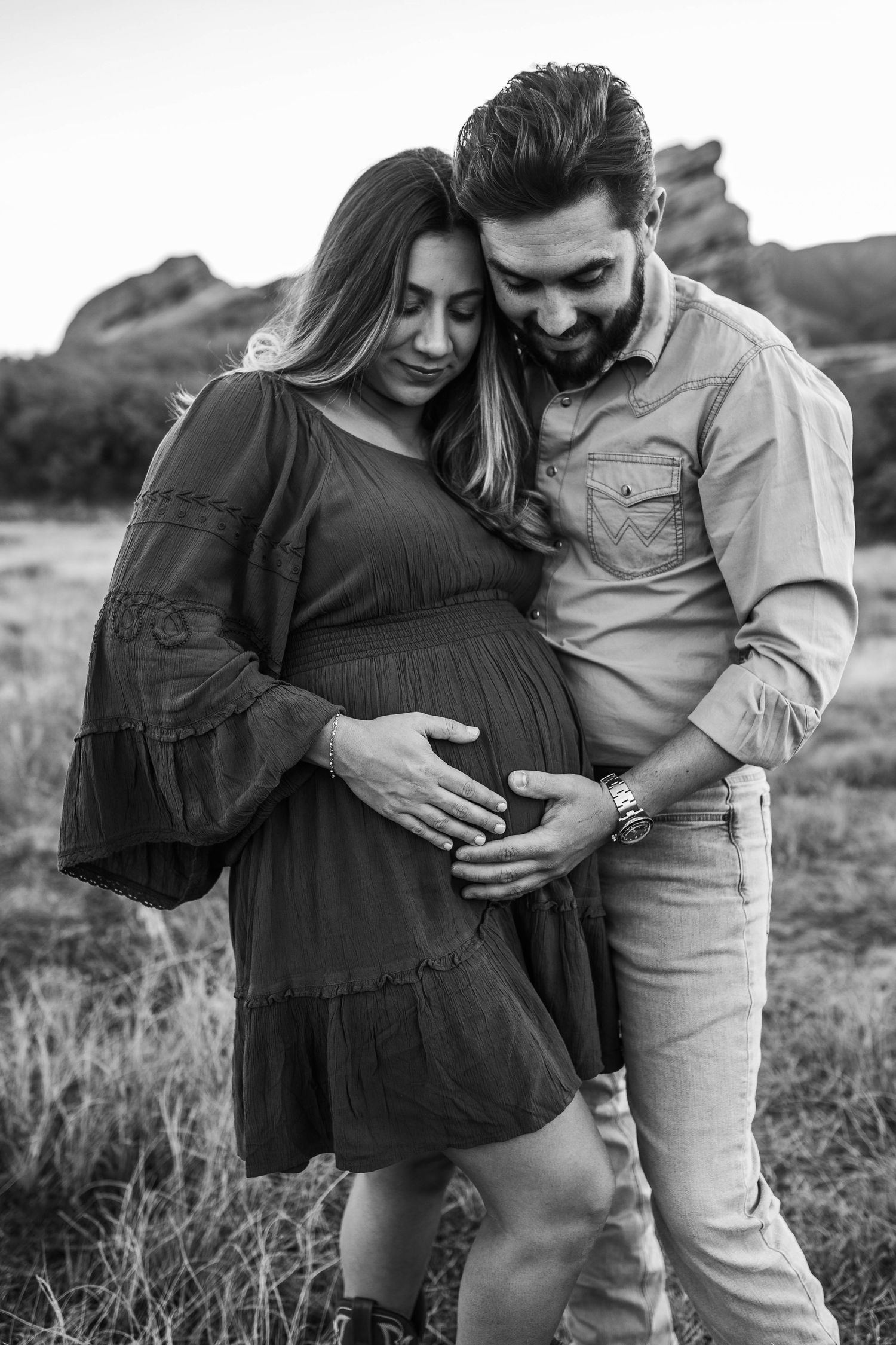 Black and white maternity photo of a couple tenderly cradling a pregnant belly outdoors near rocky landscape.