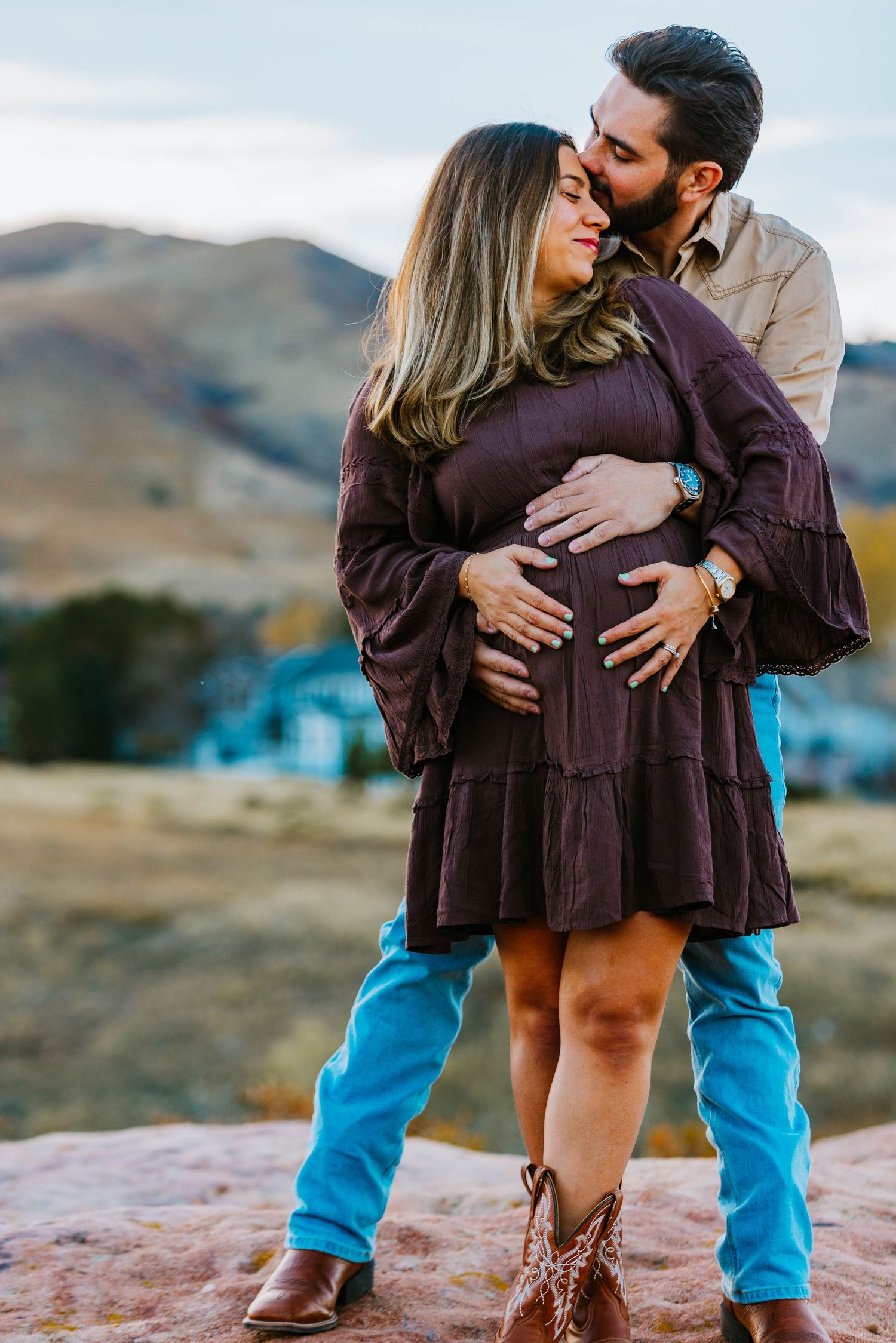 Couple sharing a tender moment outdoors, man kissing pregnant woman's cheek while cradling her belly, mountains in background.