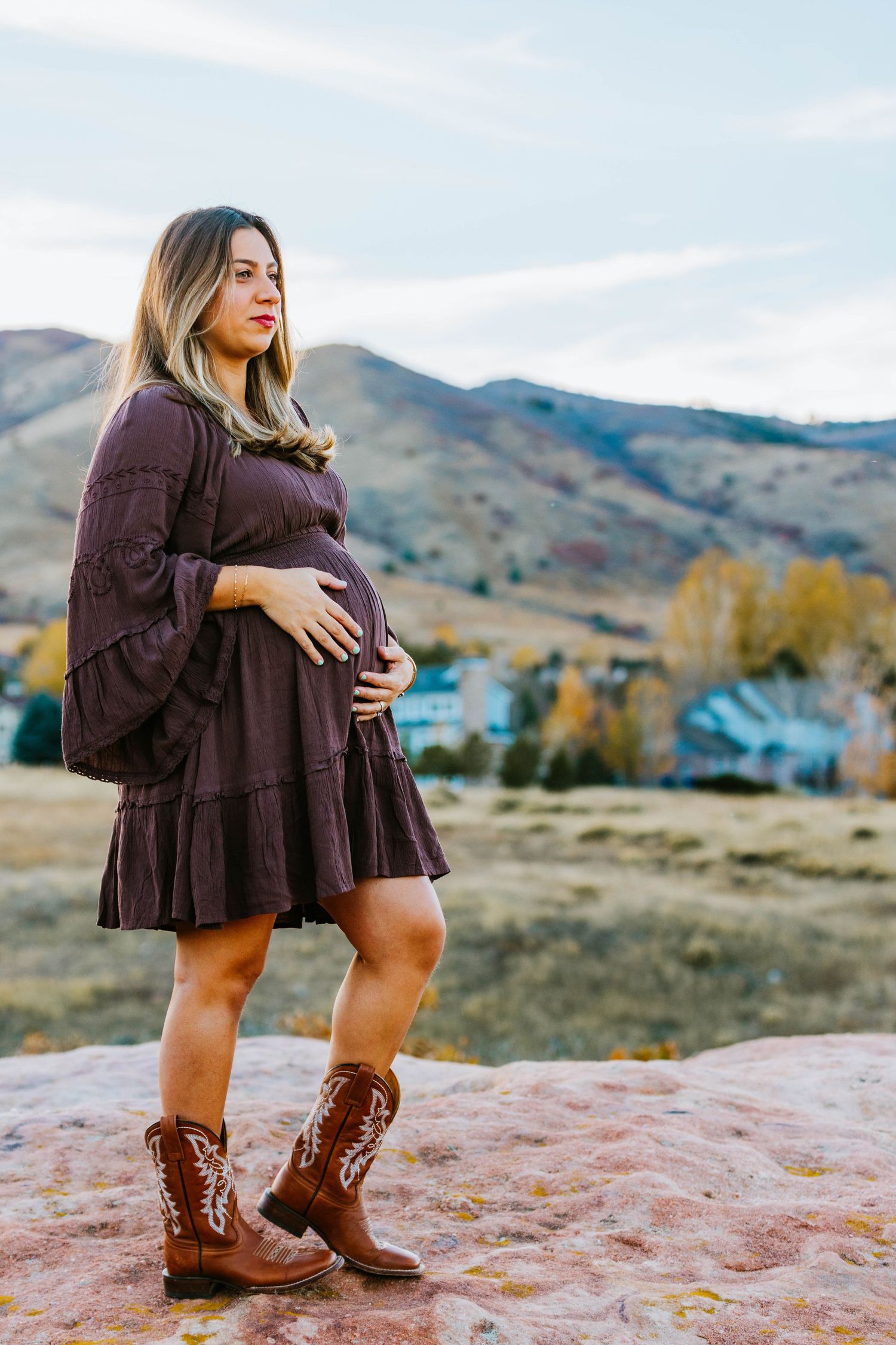 Pregnant woman in burgundy dress and cowboy boots cradling baby bump on rocky overlook with mountain backdrop.