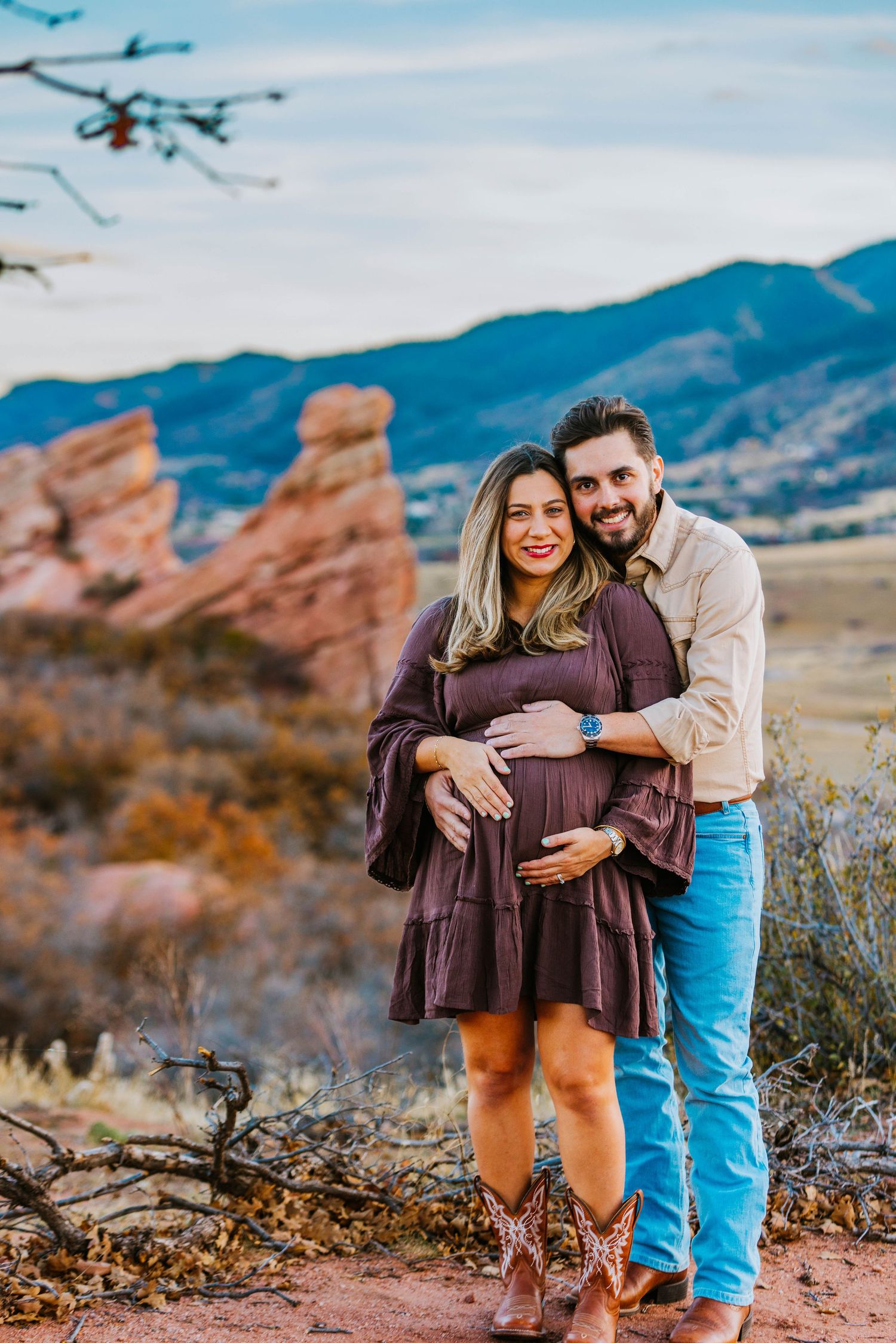 Couple poses for maternity photos at Red Rocks Colorado with mountain backdrop, woman in purple dress cradling baby bump.