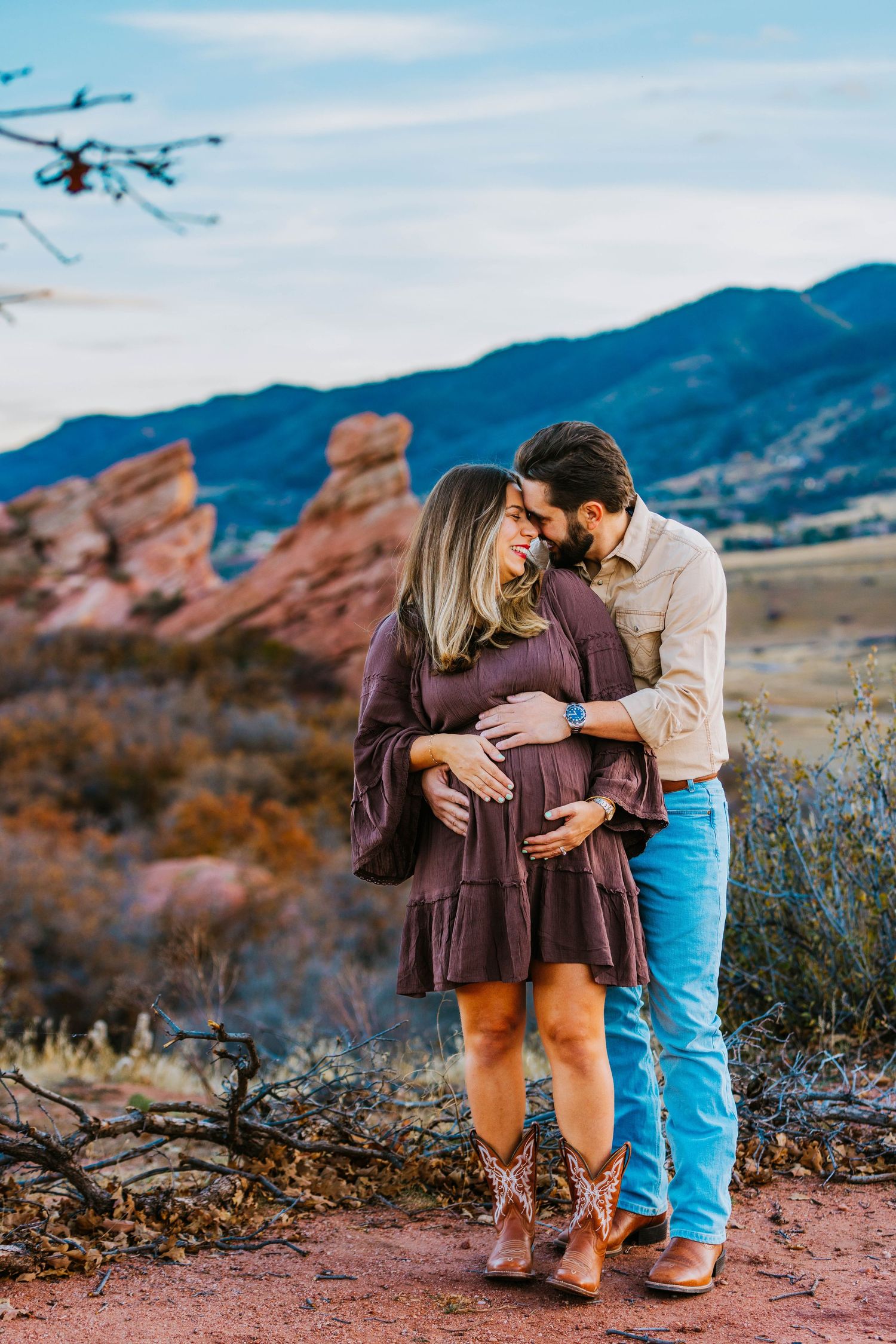 Pregnant couple embracing outdoors at Red Rocks Colorado, woman in purple dress and cowboy boots, mountains in background.