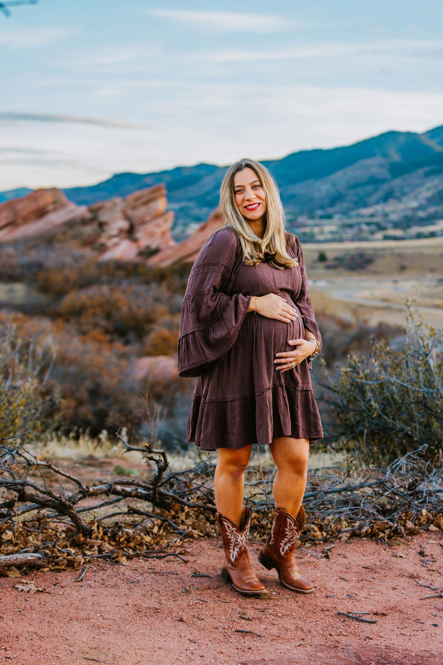 Smiling pregnant woman in brown boho dress and cowboy boots at Red Rocks Colorado in fall.