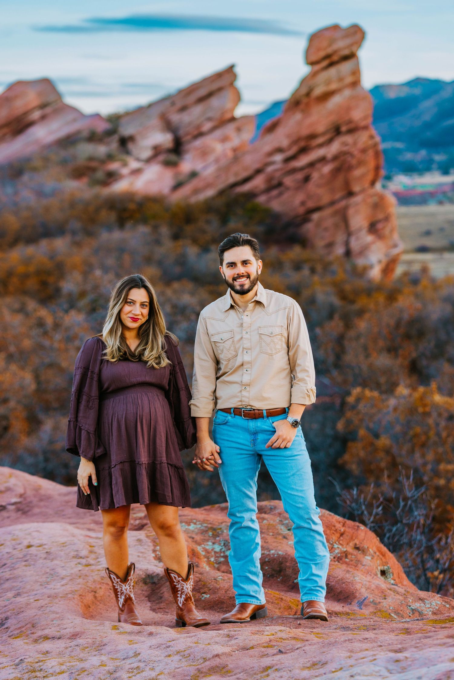 Couple holding hands at Red Rocks Park, woman in purple maternity dress with cowboy boots, man in beige shirt and blue jeans.