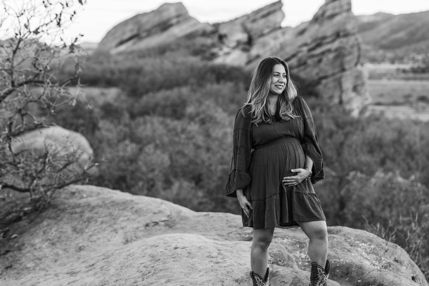 Black and white maternity photo of a smiling pregnant woman in a dress on rocks with red rock formations behind her.