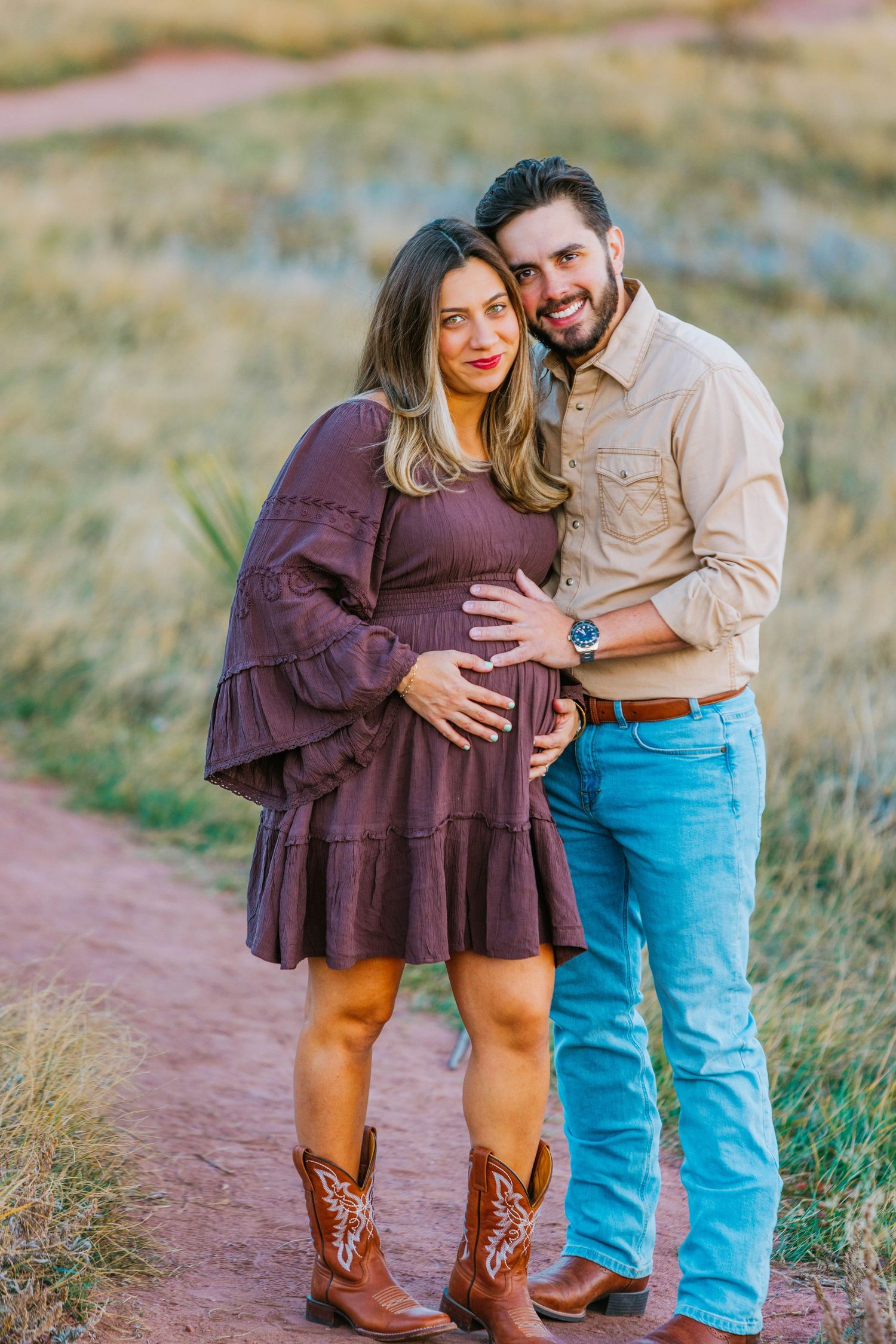 Smiling couple posing on dirt path, man embracing pregnant woman in purple dress and brown cowboy boots outdoors.