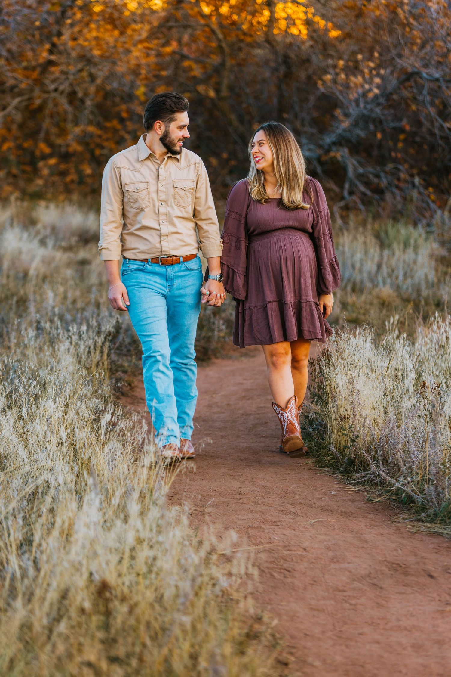 Pregnant couple holding hands walking on a dirt path surrounded by autumn foliage in a scenic outdoor setting.