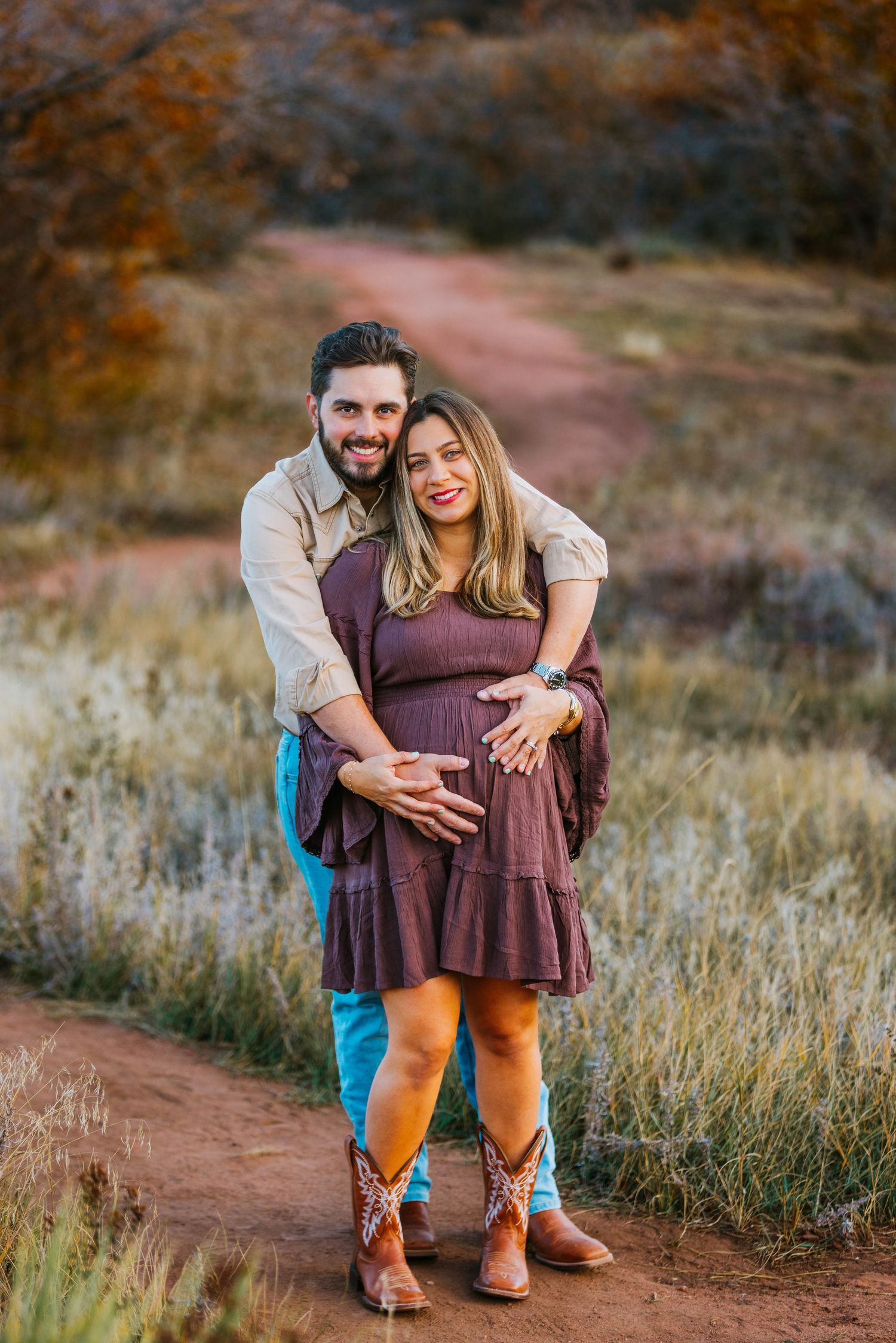 Happy couple posing on red dirt path, pregnant woman in purple dress and cowboy boots embraced by man in blue jeans.