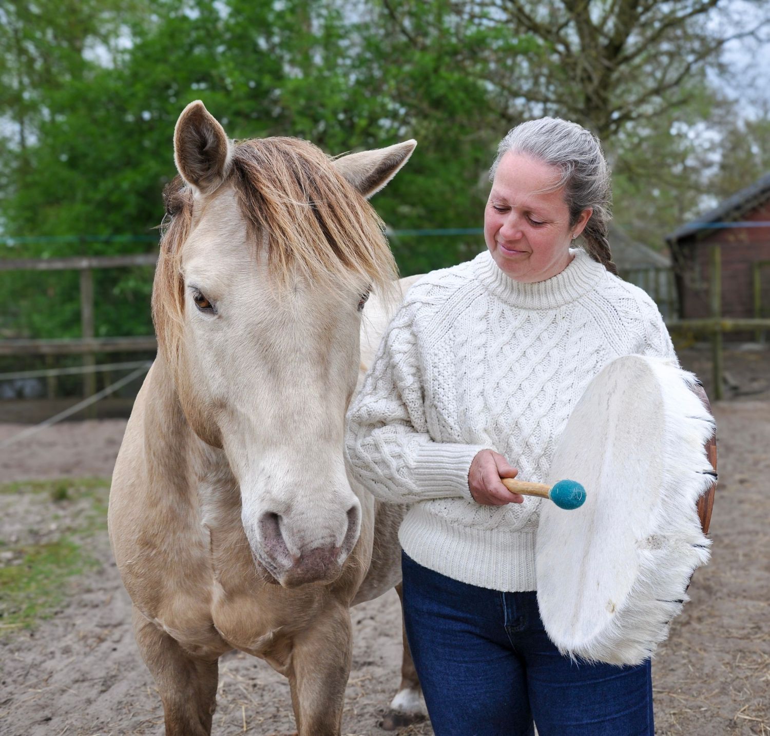 Woman smiling while holding a frame drum and drumstick next to a palomino horse outdoors.