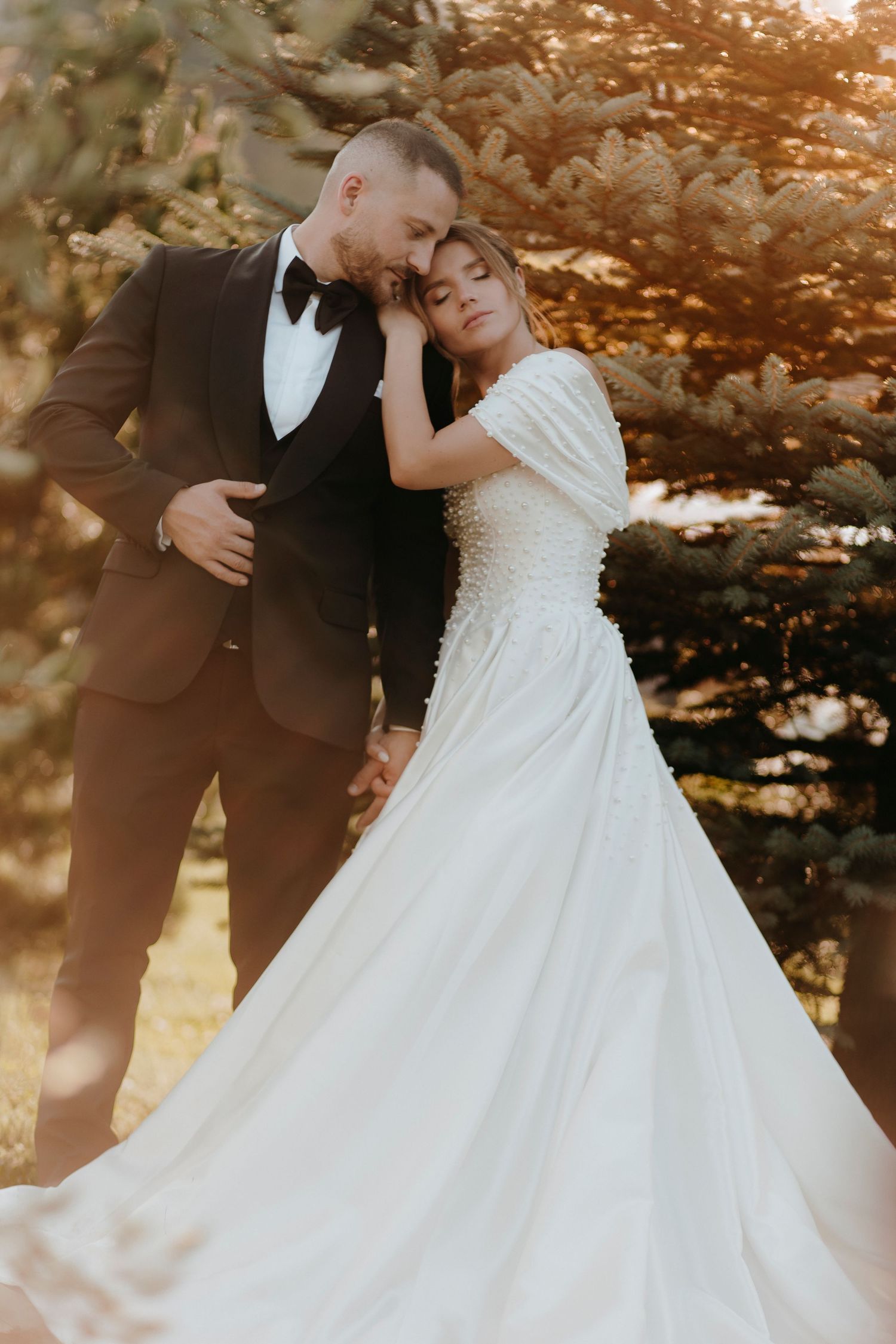 A bride in a flowing white gown leans on her groom in a black tuxedo amid golden sunlit trees.