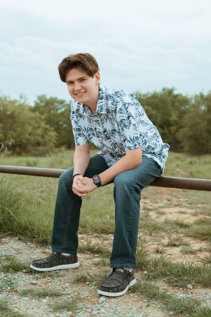 Teen boy in blue leaf-print shirt and jeans sitting on a metal rail smiling outdoors.