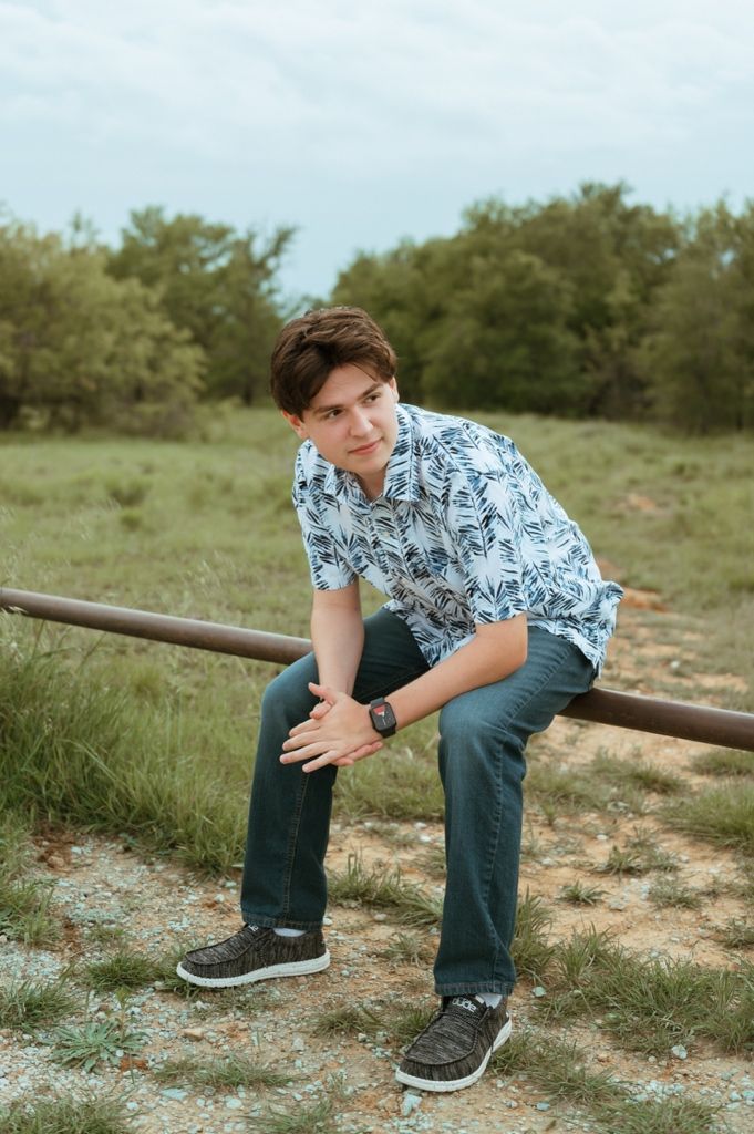 Teen boy in floral shirt and jeans sits on a metal rail in a grassy outdoor field.