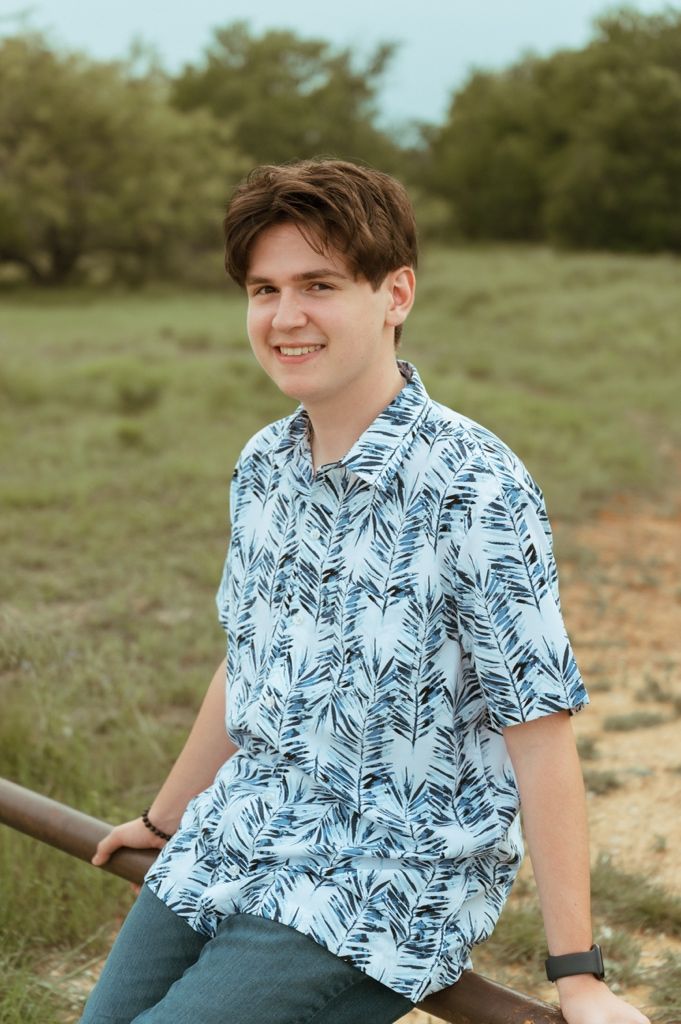 Young man in blue tropical shirt leaning on a metal rail smiling in a grassy field.