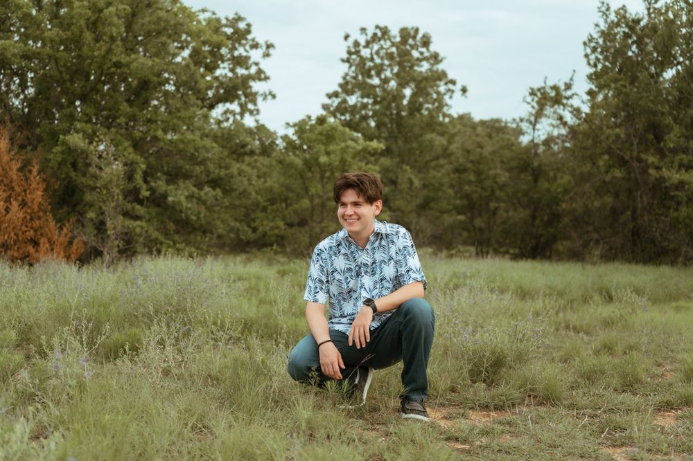 Teenage boy in blue floral shirt crouching in a grassy outdoor field surrounded by trees.