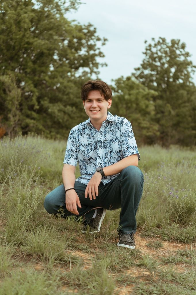 Smiling young man in blue leaf-print shirt crouching in a grassy field with trees in the background.