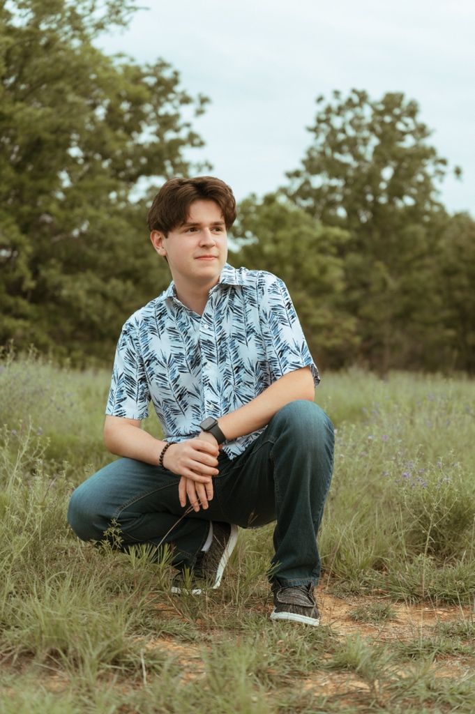 Teen boy in blue leaf-print shirt crouching in grassy field with trees behind him.