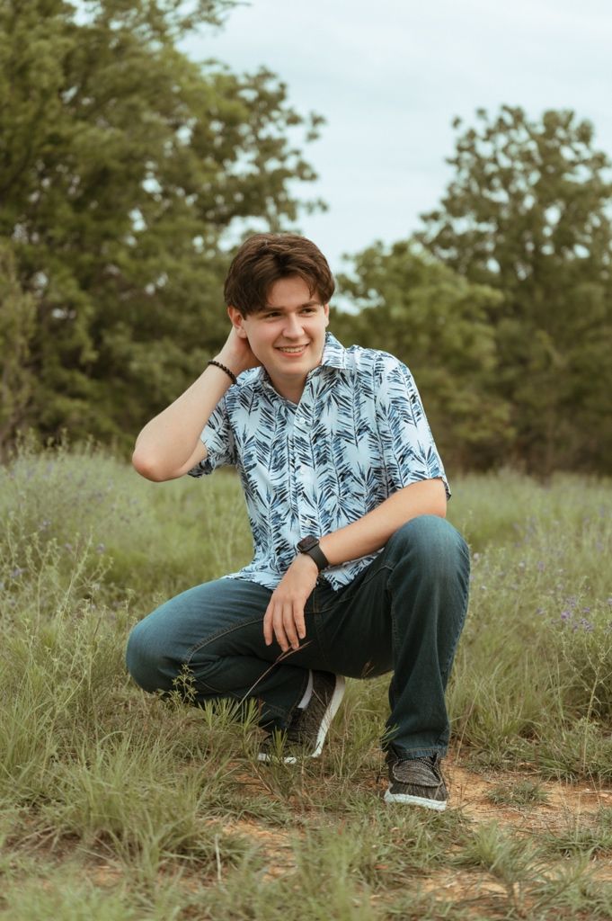 Young man in blue floral shirt crouching in a wildflower meadow with trees in background.