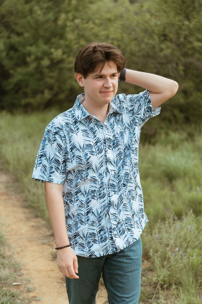 Young man in a blue tropical print shirt poses on a dirt path surrounded by lush green trees.
