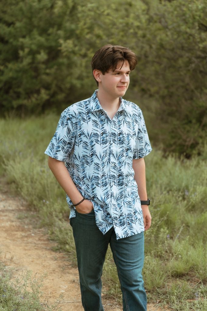 Young man in blue tropical print shirt standing on a nature trail surrounded by green foliage.