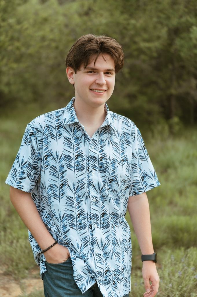 Smiling teenage boy in blue floral shirt standing outdoors in a green natural setting.