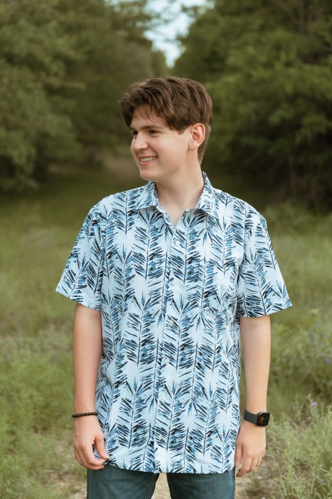 Smiling teen in a blue leaf-print button-up shirt posing outdoors in a green meadow setting.
