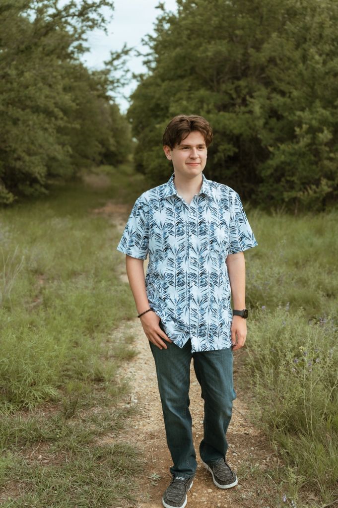 Young man in blue floral shirt and teal pants standing on a nature trail surrounded by green trees.