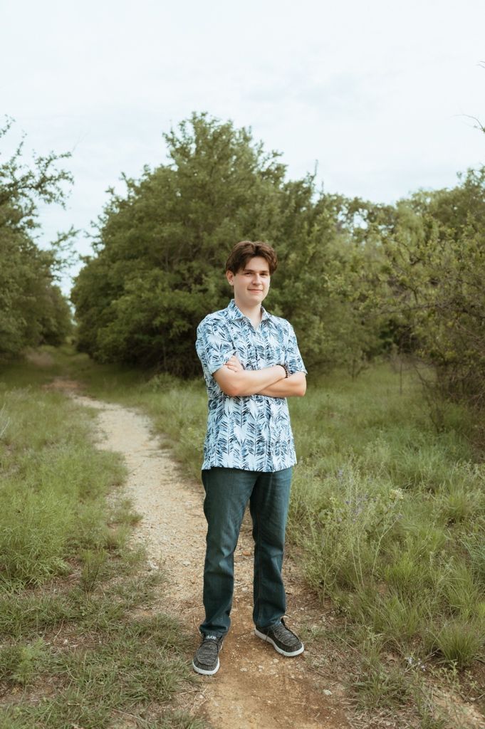 Teen boy in patterned blue shirt standing with arms crossed on a dirt path in a lush green landscape.