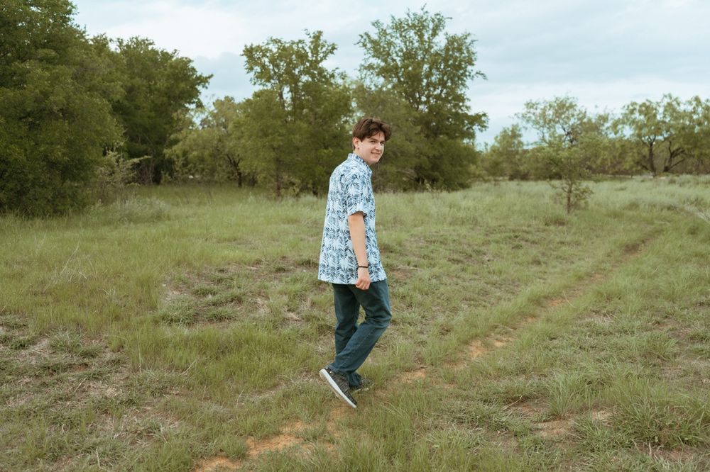 Young man in blue floral shirt walking through green meadow with trees in background.