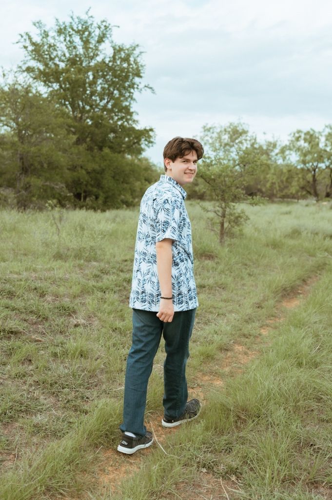Young man in floral shirt standing on a grassy trail smiling over shoulder.
