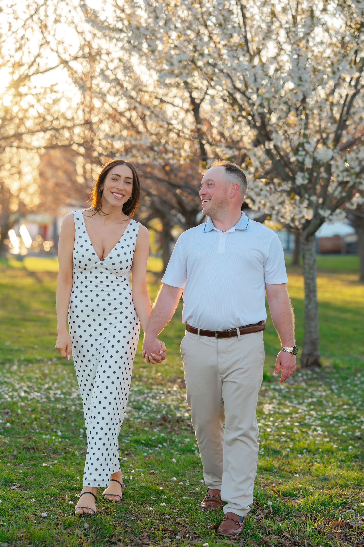 Couple holding hands walking through a blooming cherry blossom park at golden hour, woman in polka dot jumpsuit.