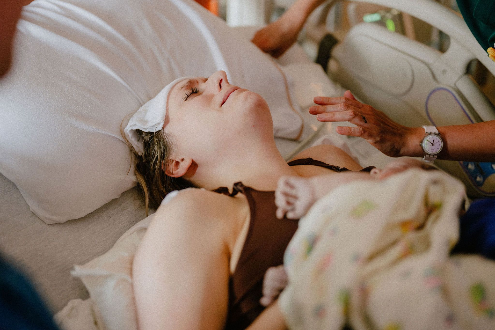 Exhausted new mother lying in hospital bed smiling after childbirth, holding newborn baby wrapped in blanket.