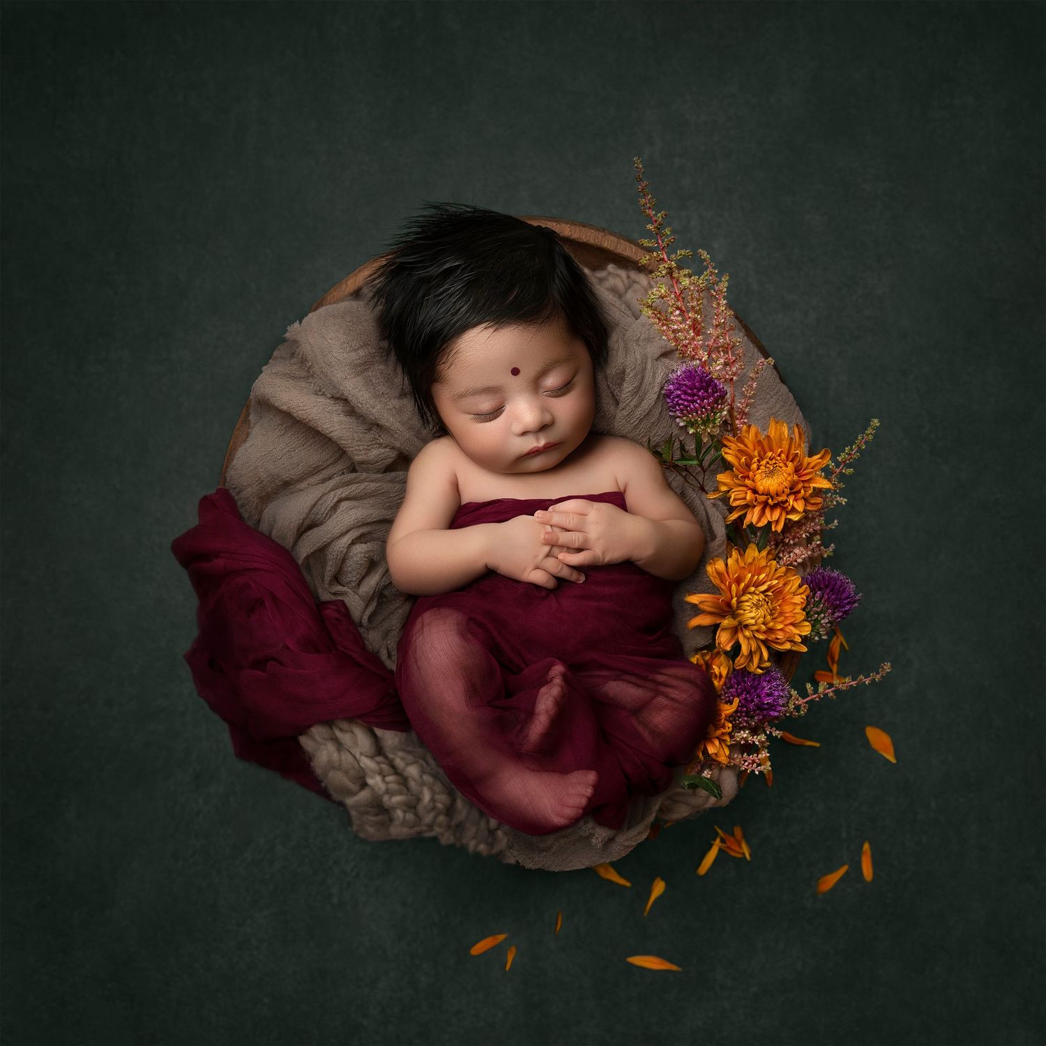 Newborn baby sleeping peacefully in a wooden bowl with red wrap and colorful flowers on green background.