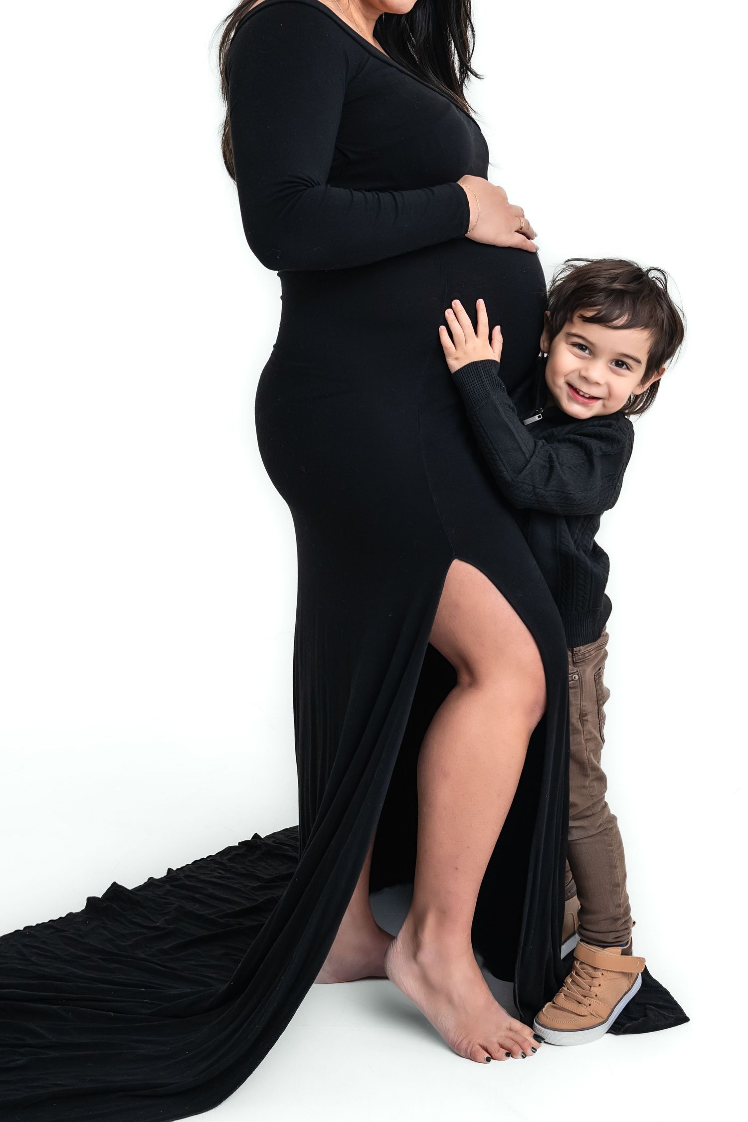 Pregnant woman in black gown hugged by smiling toddler boy in maternity photo session.