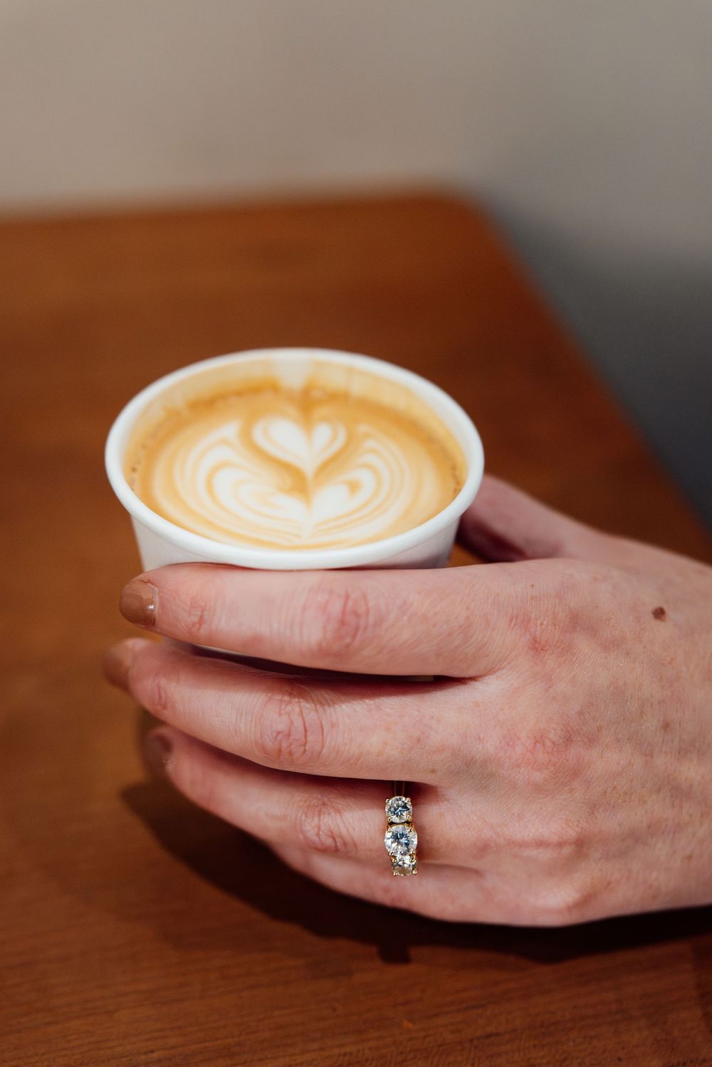 A hand wearing a diamond ring holds a white paper cup with heart latte art on a wooden table.