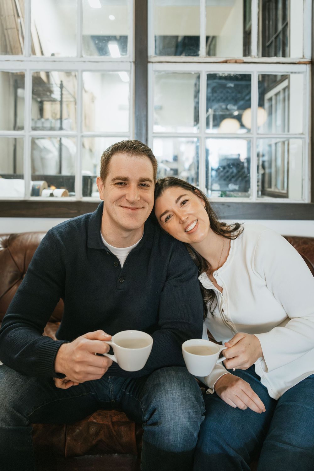 A smiling couple sitting on a leather sofa holding coffee cups in a cozy cafe setting.