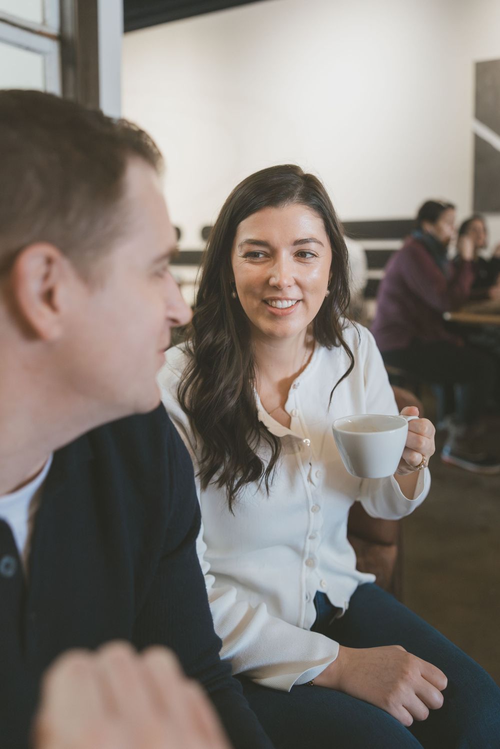 A smiling woman holding a white coffee cup chats with a man in a cozy cafe setting.