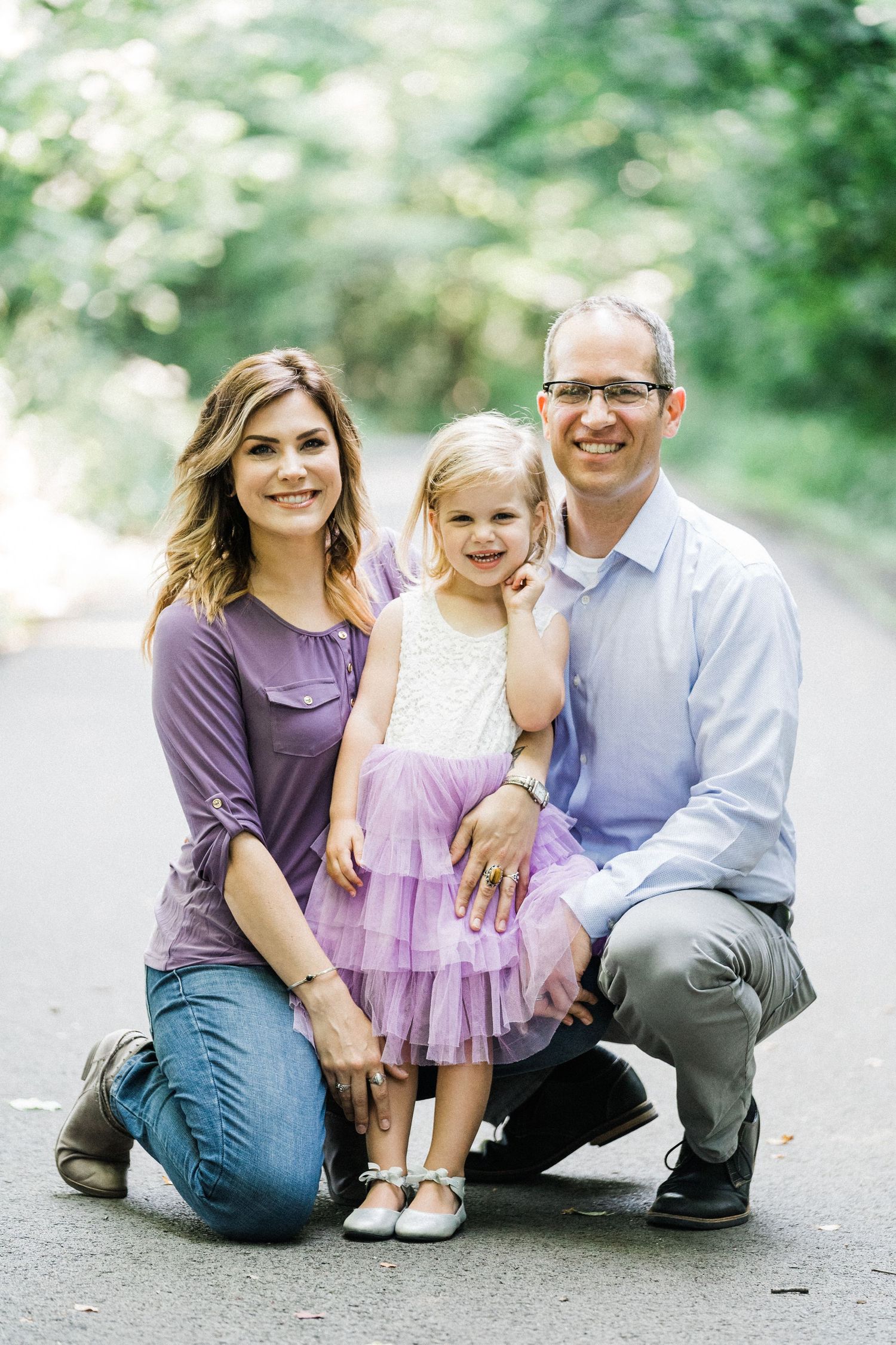A smiling family of three poses outdoors, with parents kneeling beside their young daughter in a purple tutu dress.
