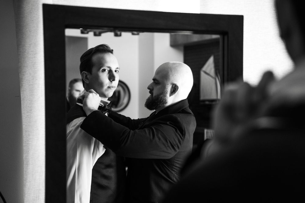 Groomsman helping groom adjust his tie while looking in a mirror in black and white photo.