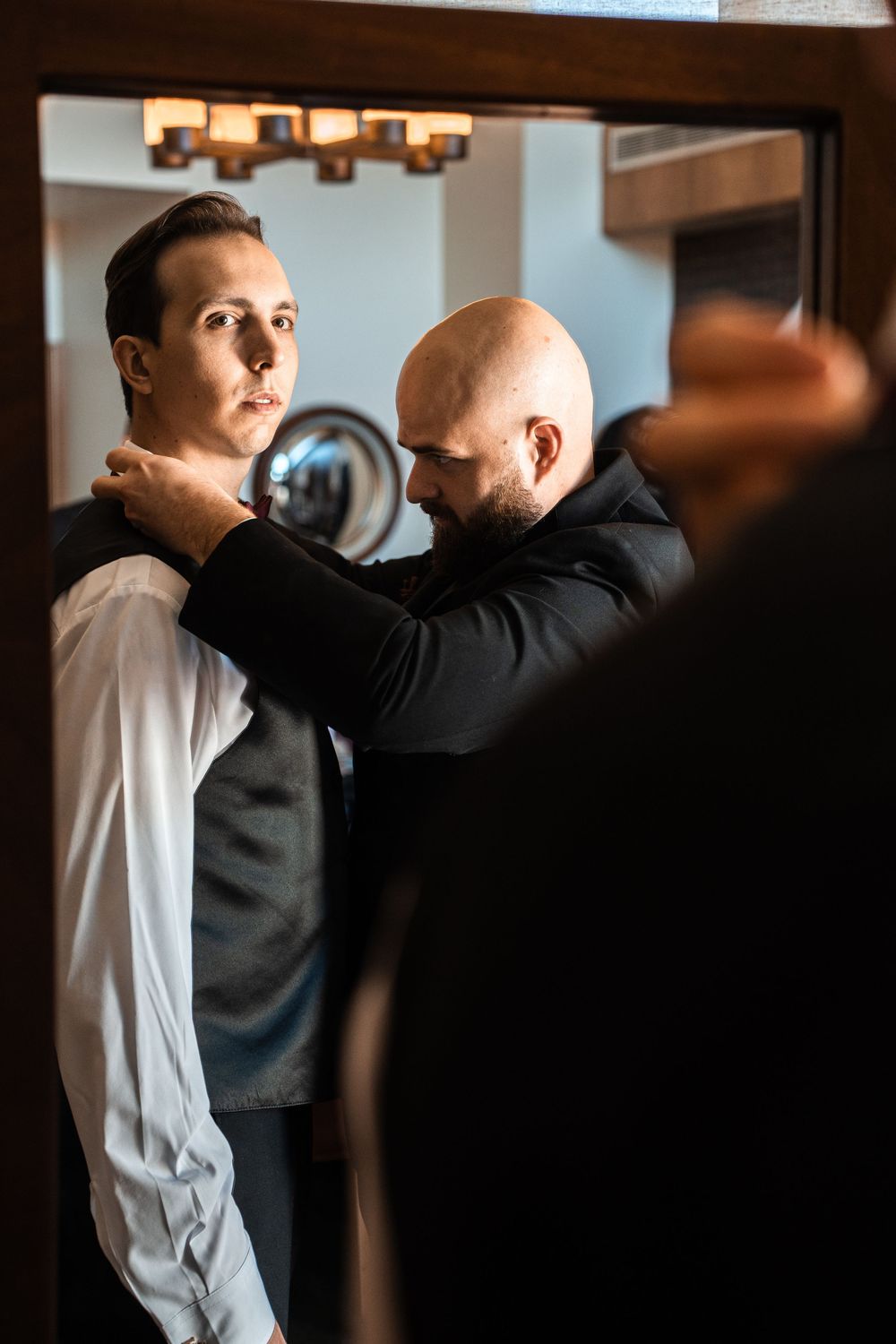 A bald man helps another man adjust his collar in front of a mirror in a dimly lit room.
