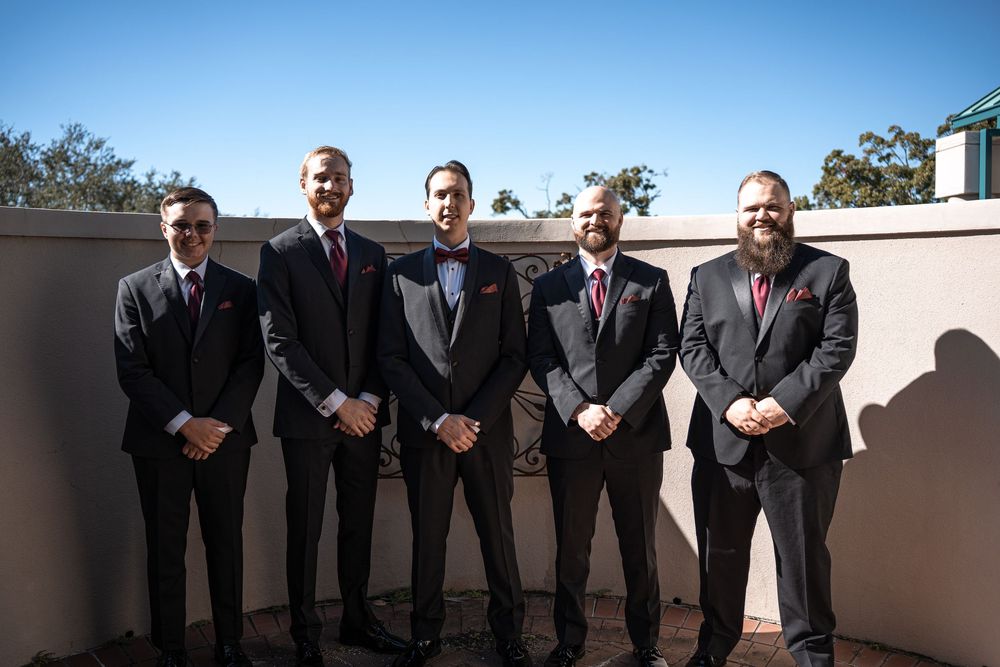 Five groomsmen in dark suits with burgundy ties pose outdoors on a sunny day at a wedding venue.