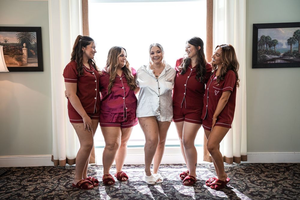 Bride in white pajamas poses with five bridesmaids in matching red pajama sets, laughing together by a bright window.
