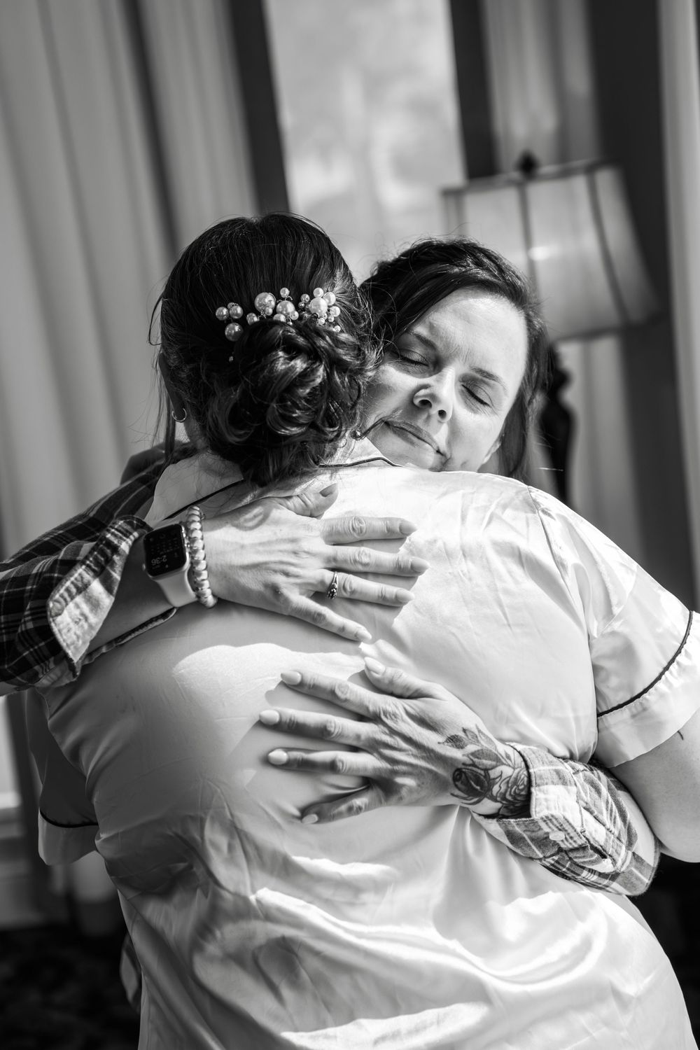Black and white photo of a bride in a white robe sharing an emotional hug with a woman on her wedding day.