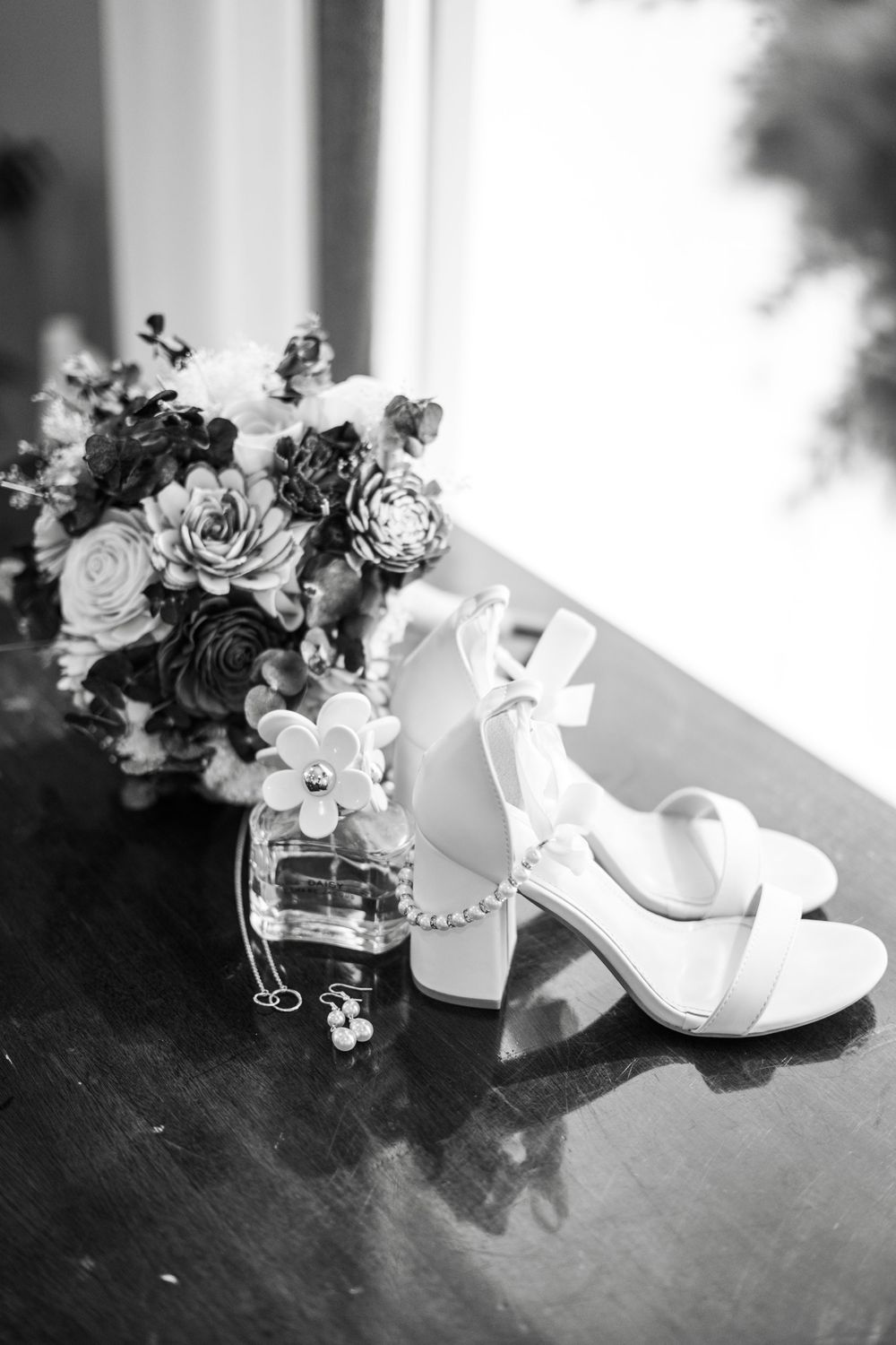 Black and white photo of bridal heels, floral bouquet, perfume bottle, and pearl necklace on a wooden surface.
