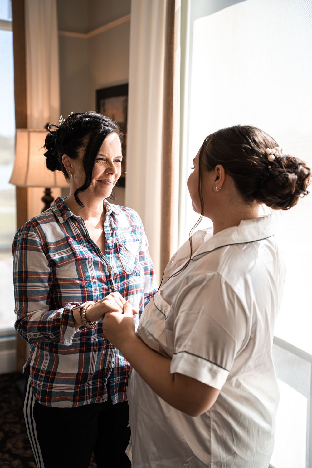 Two women in casual attire hold hands and smile near a bright window during wedding preparations.