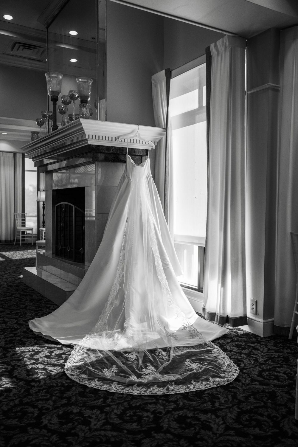 Elegant white wedding dress with lace train hanging near a fireplace in a black and white photo.