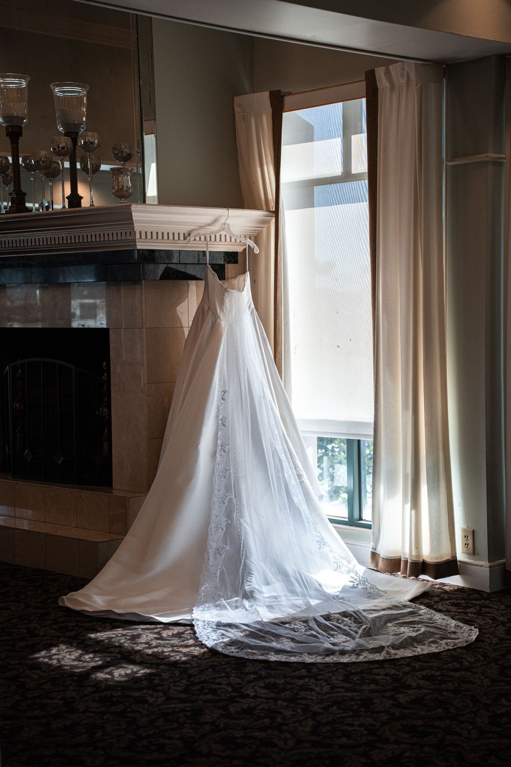 Elegant white wedding dress with lace train hanging near a fireplace and window in a classic bridal suite.