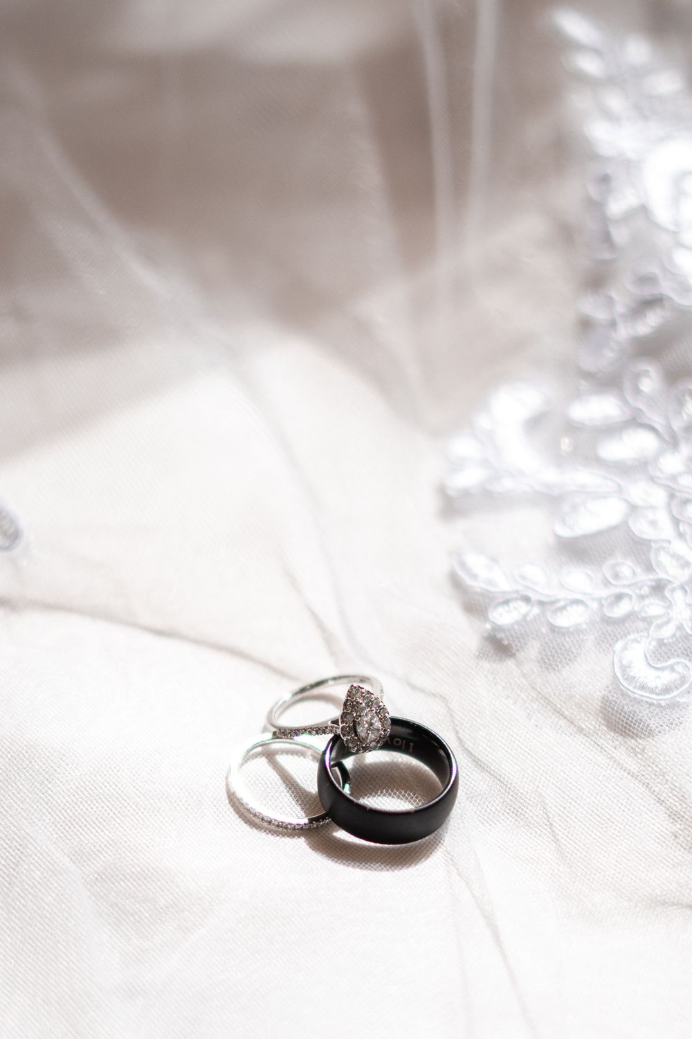 Wedding rings set on white bridal veil, featuring diamond engagement ring, eternity band, and black tungsten band.