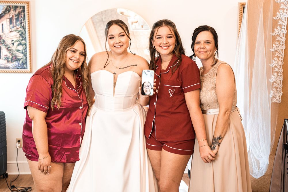 Bride in white gown poses with three women in maroon and beige outfits during wedding preparations indoors.